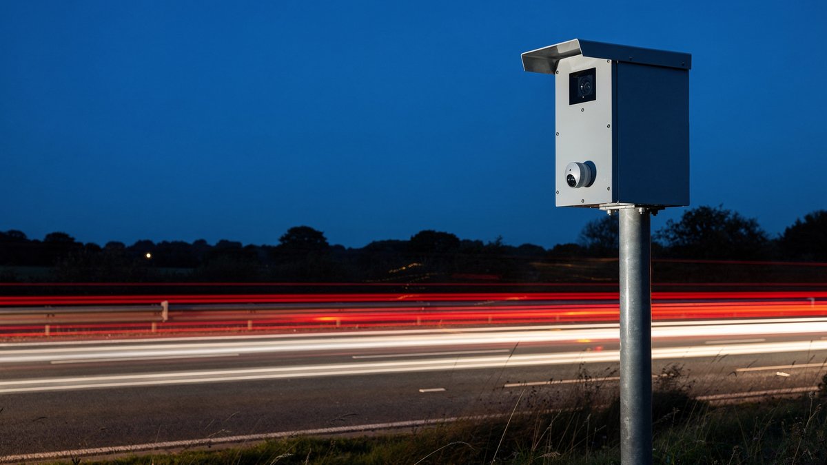 A roadside traffic camera on a grey pole at dusk, with red and white light trails from passing traffic behind it